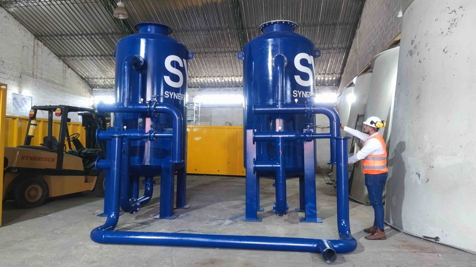 Técnico inspeccionando una planta de tratamiento de aguas residuales modular de color verde marca BIOMAX SYNERTECH, que incluye módulos de pretratamiento, decantación, DAF y oxidación avanzada. La planta está ubicada en un entorno industrial con techo alto y tuberías visibles. - ver más información