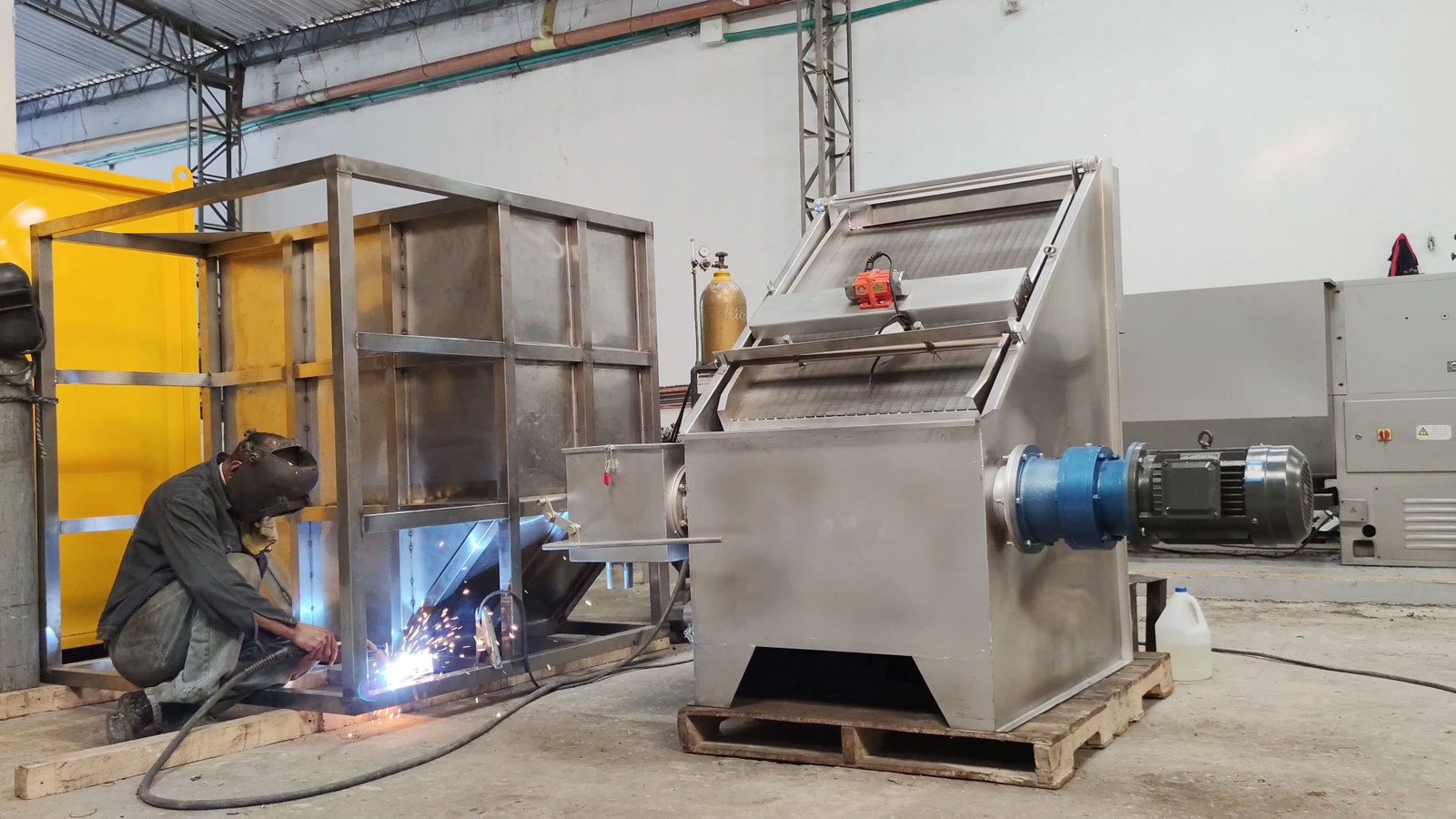 Trabajador soldando un tamiz estático con tornillo prensa en un entorno industrial, usando equipo de protección. Se ven chispas de soldadura y, al fondo, otras máquinas, gabinetes eléctricos y conductos de ventilación. - ver mas información