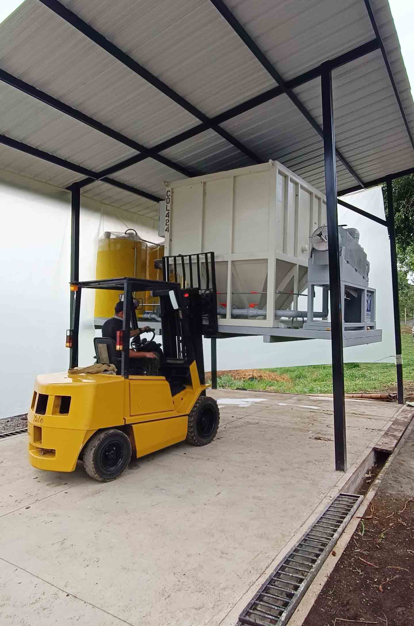 Instalación de un filtro industrial grande marcado con 'SYNERTECH' en un sitio de construcción. Una excavadora con el cubo levantado parece ayudar en el posicionamiento del filtro. Un trabajador con camiseta naranja de alta visibilidad y casco supervisa el proceso. Al fondo, se observa una estructura que parece una torre industrial, parte de la infraestructura del sitio.